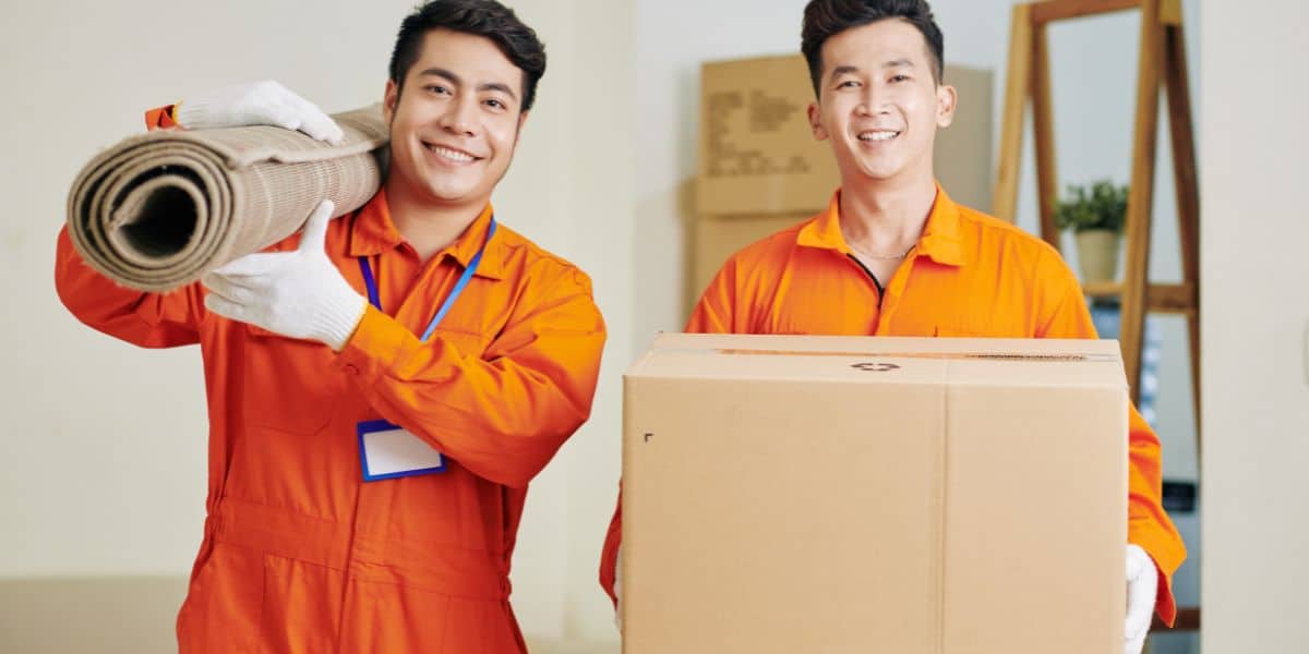 Two movers in orange uniforms smiling, one carrying a rolled-up rug and the other holding a cardboard box.