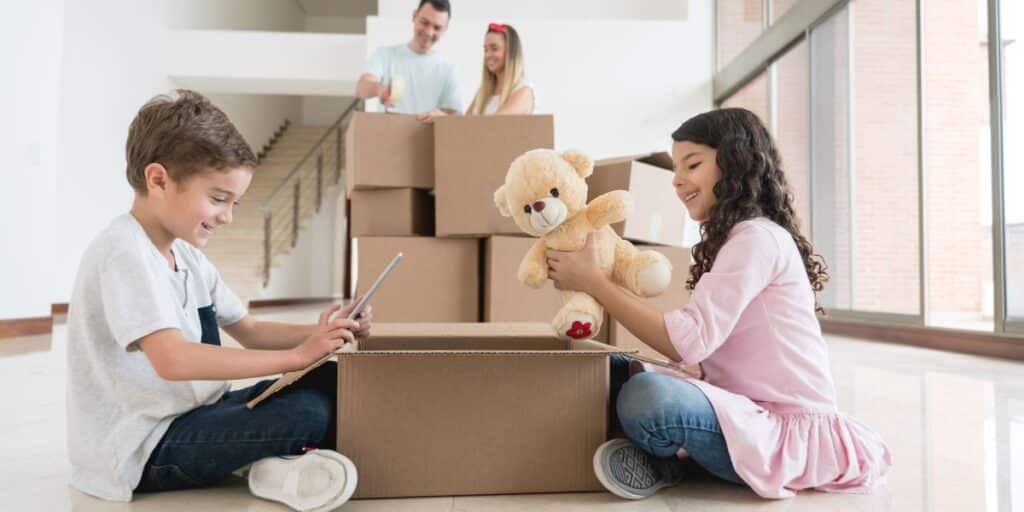 Two children helping pack moving boxes while their parents organize in the background. One child is holding a tablet, and the other is holding a teddy bear, creating a positive family atmosphere during the move.