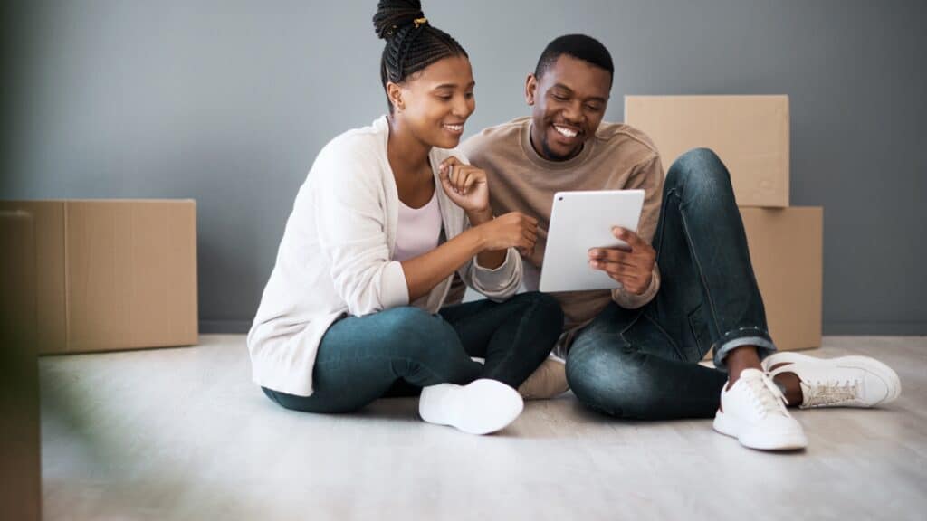 Smiling couple sitting on the floor with moving boxes, reviewing their moving supplies checklist on a tablet.