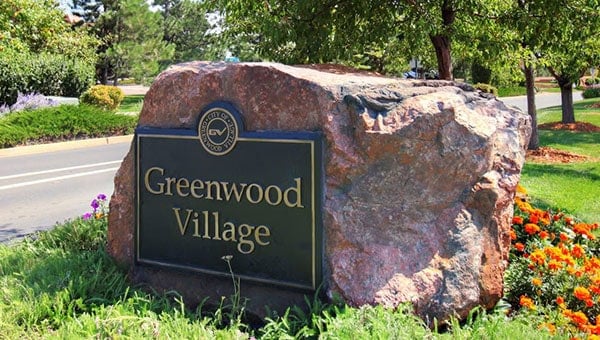 Greenwood Village entrance sign on a large rock with colorful flowers and trees in the background.