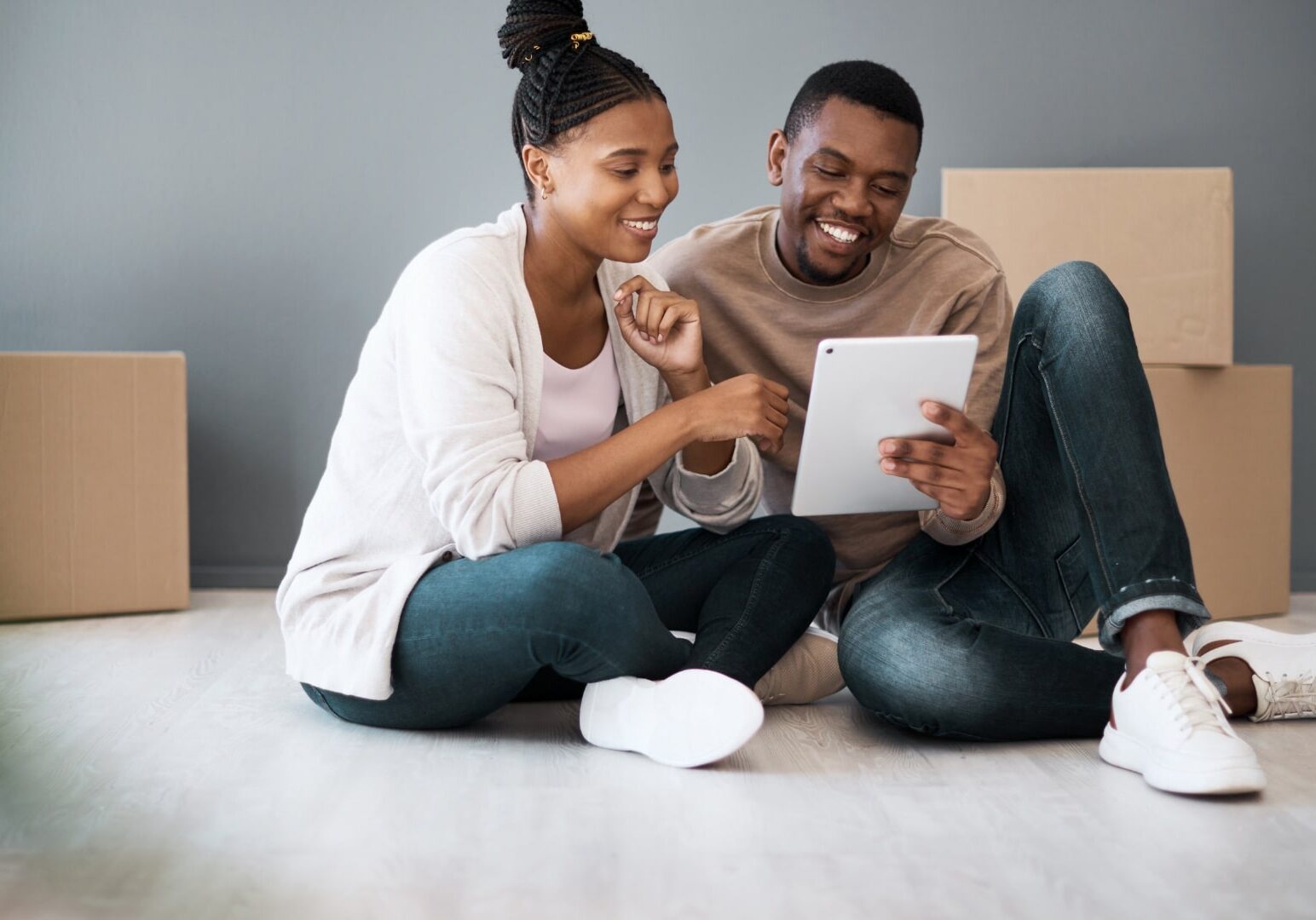 Smiling couple sitting on the floor with moving boxes, reviewing their moving supplies checklist on a tablet.